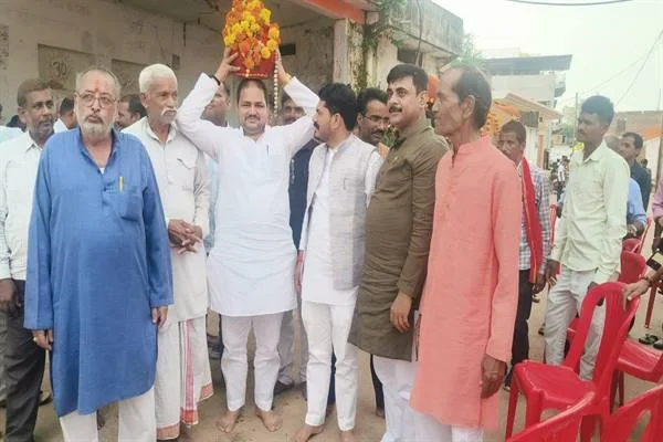 ग्रामीण मार्ग पर खड़े लोगों के बीच सफेद कुर्ता-पायजामा पहने एक व्यक्ति सिर पर फूलों से सजी वस्तु लिए हुए