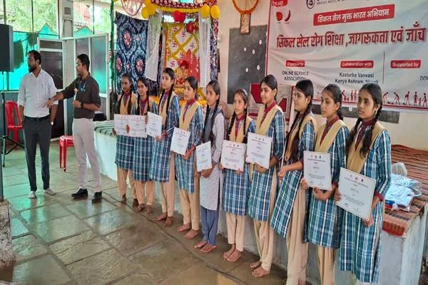 प्रमाण-पत्र लिए कतार में खड़ी छात्राएं और उनके पास खड़े दो पुरुष, पीछे सिकल सेल जागरूकता शिविर का बैनर लगा है
