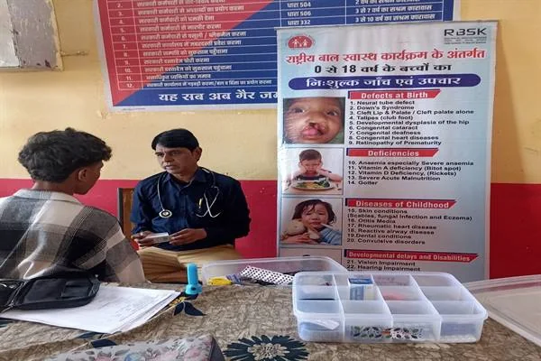 मेज के पास बैठे स्वास्थ्यकर्मी से बात करता एक छात्र, साथ में जांच सामग्री और बाल स्वास्थ्य संबंधी पोस्टर