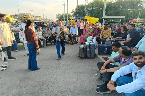 सड़क पर दो महिला कलाकारों के सामने कुर्सियों पर बैठे लोग नुक्कड़ नाटक देखते हुए